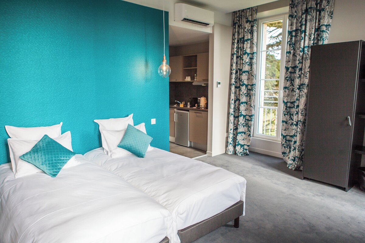 Een slaapkamer met een groot bed, twee kussens, een wand in teal, een kledingkast en uitzicht op de keuken via een geopende deur.
