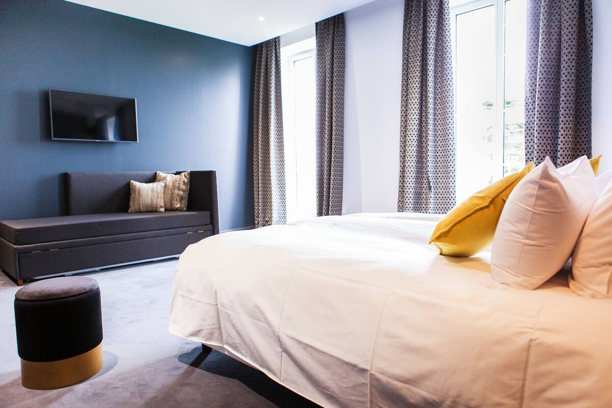 Une chambre moderne avec un grand lit, un canapé foncé, un téléviseur à écran plat et une fenêtre avec des rideaux.