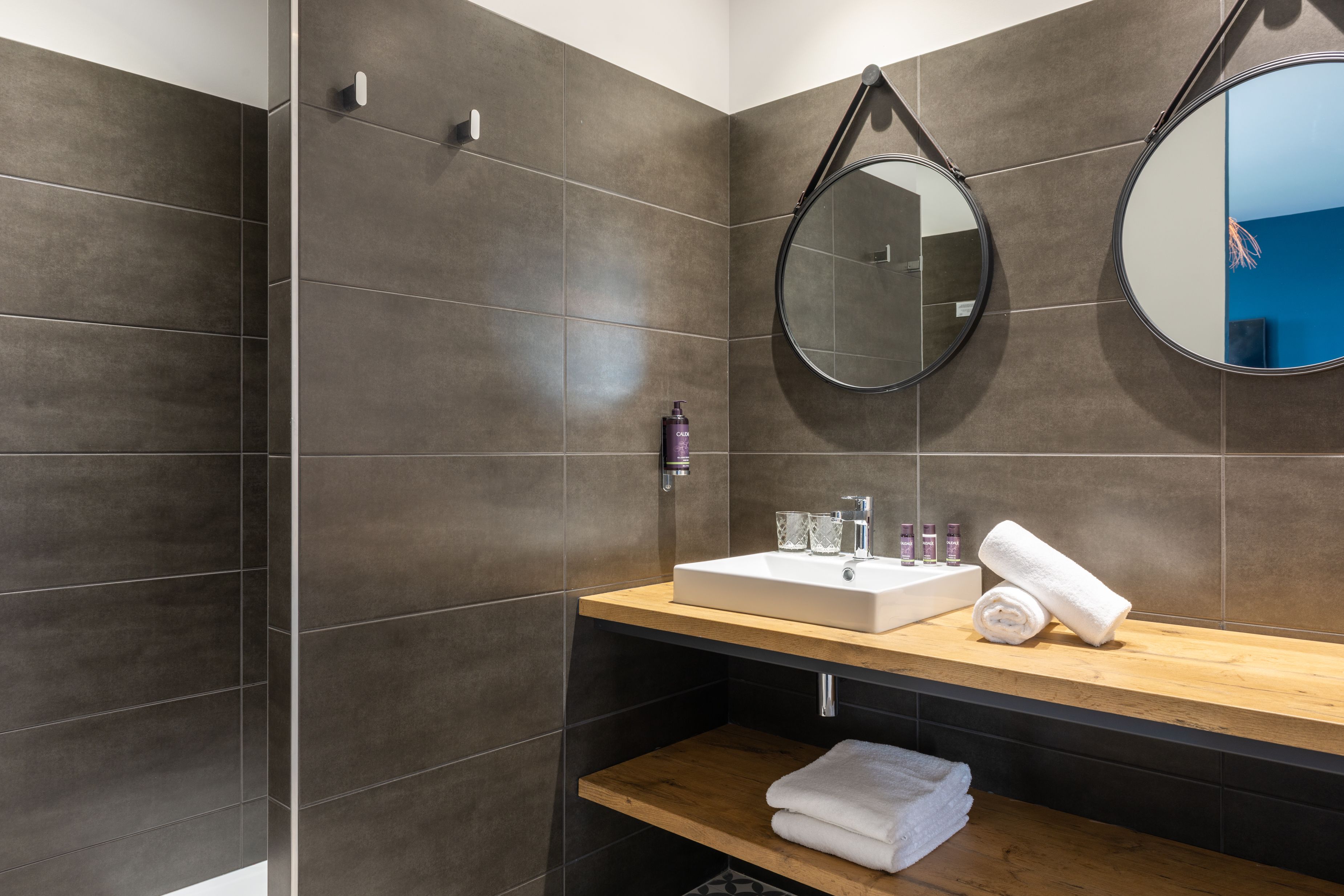 A modern bathroom with dark tiles, a wooden vanity, a sink, a mirror, and a shower area.