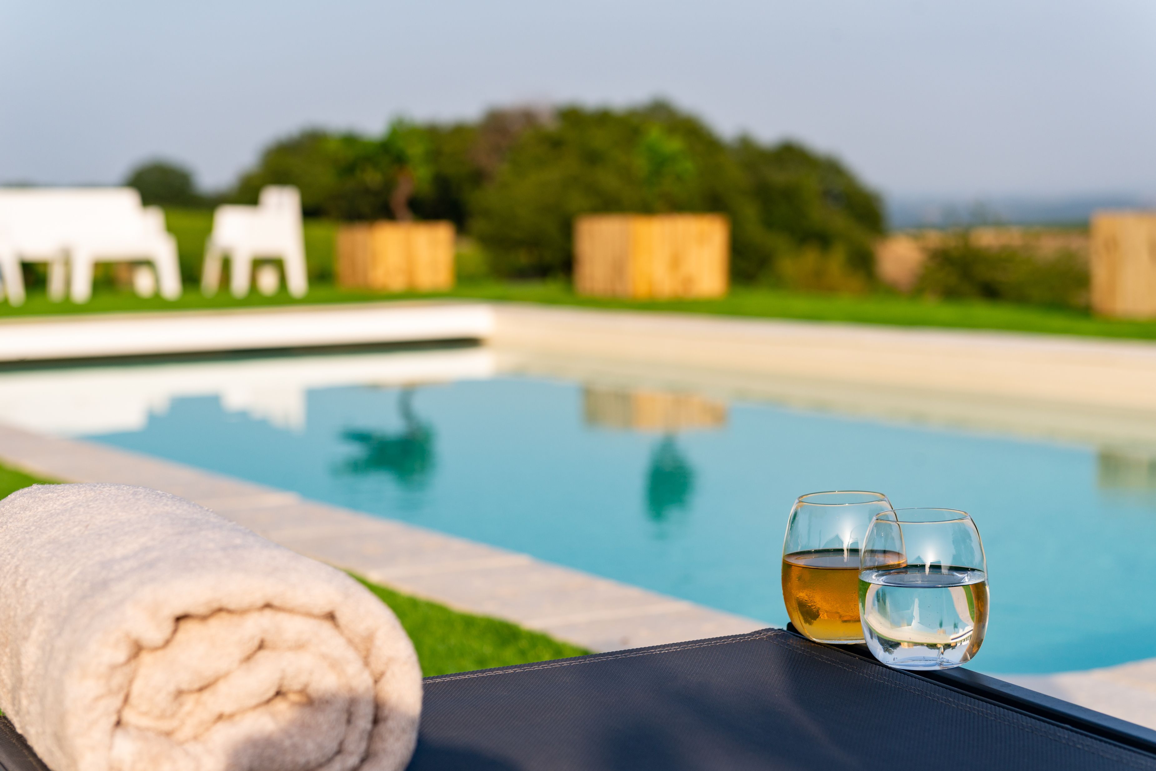 Escena junto a la piscina, con una toalla y dos vasos de agua, cielo despejado y follaje en el fondo.