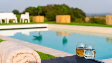 Escena junto a la piscina, con una toalla y dos vasos de agua, cielo despejado y follaje en el fondo.