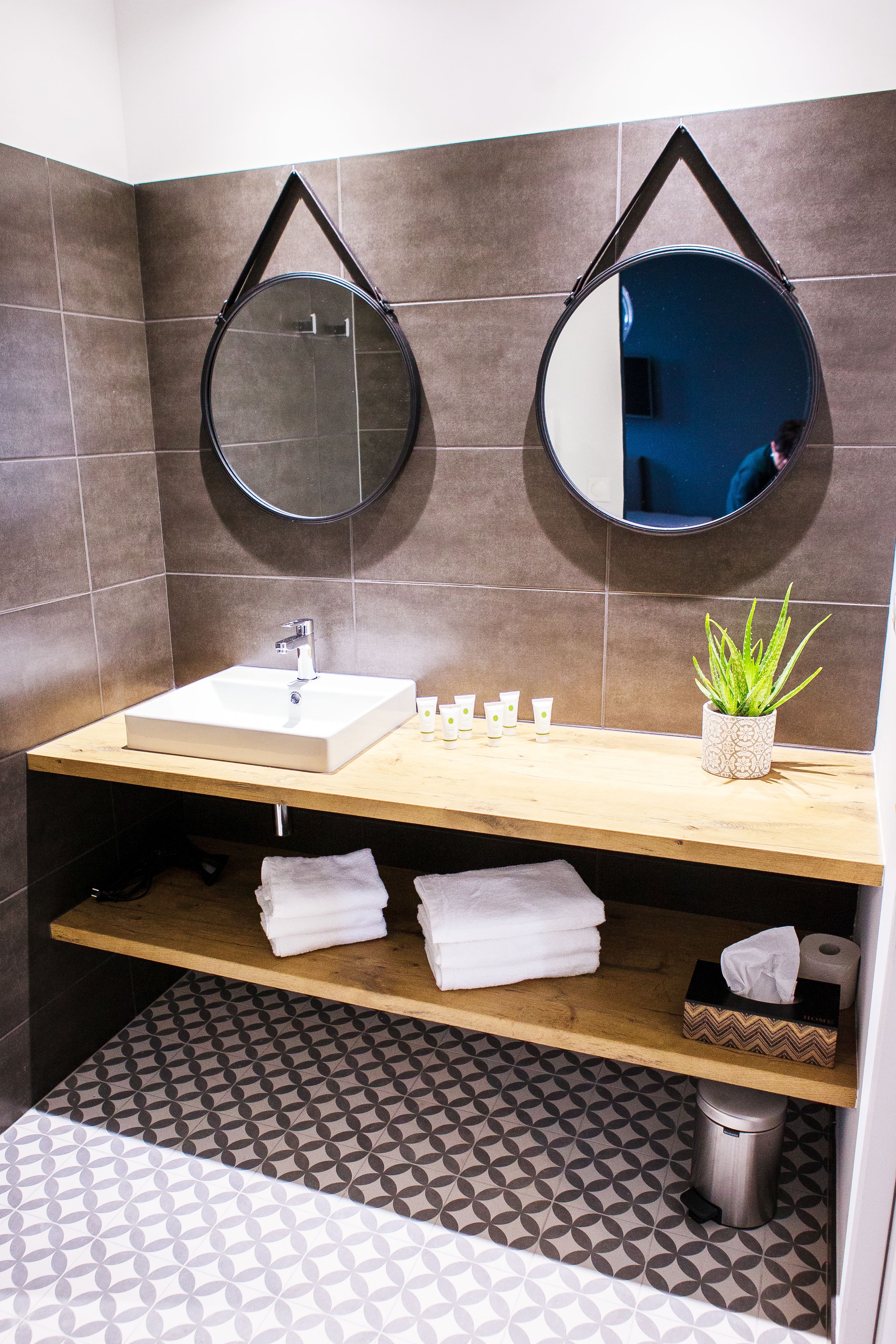 A bathroom with a white sink, two round mirrors, a potted plant, and a shelf with towels.