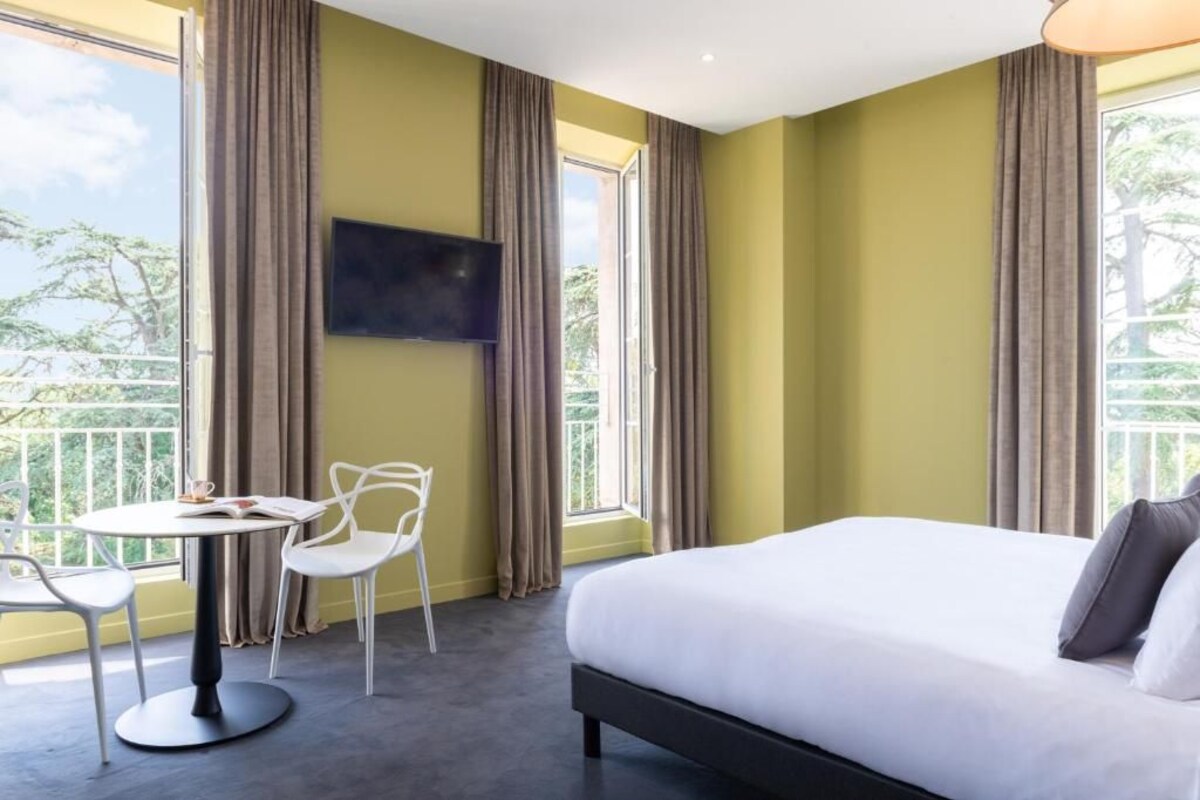 Een hotelkamer met een bed, een aan de muur bevestigde televisie, een eettafel met stoelen en grote ramen met gordijnen.