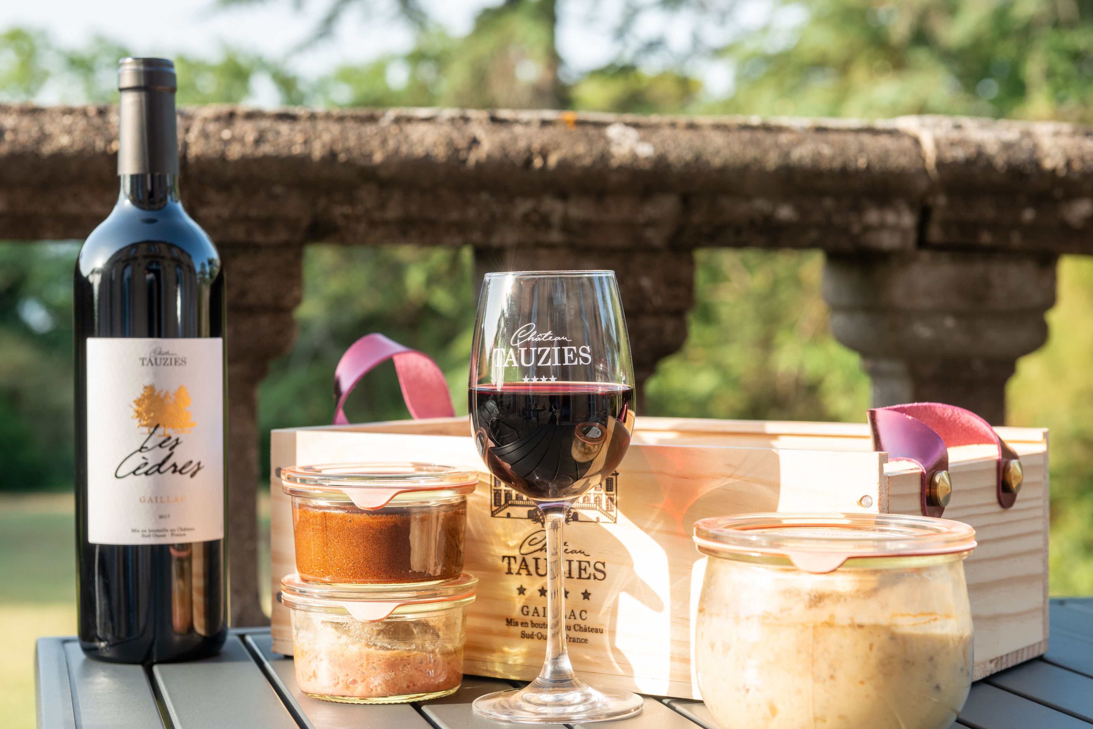 Una botella de vino, una copa de vino y dos frascos de comida sobre una mesa al aire libre.