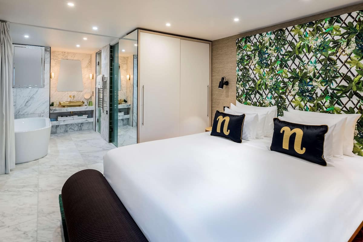 A modern hotel room with a large bed, a bathroom with a bathtub, and a glass door leading to a shower area.
