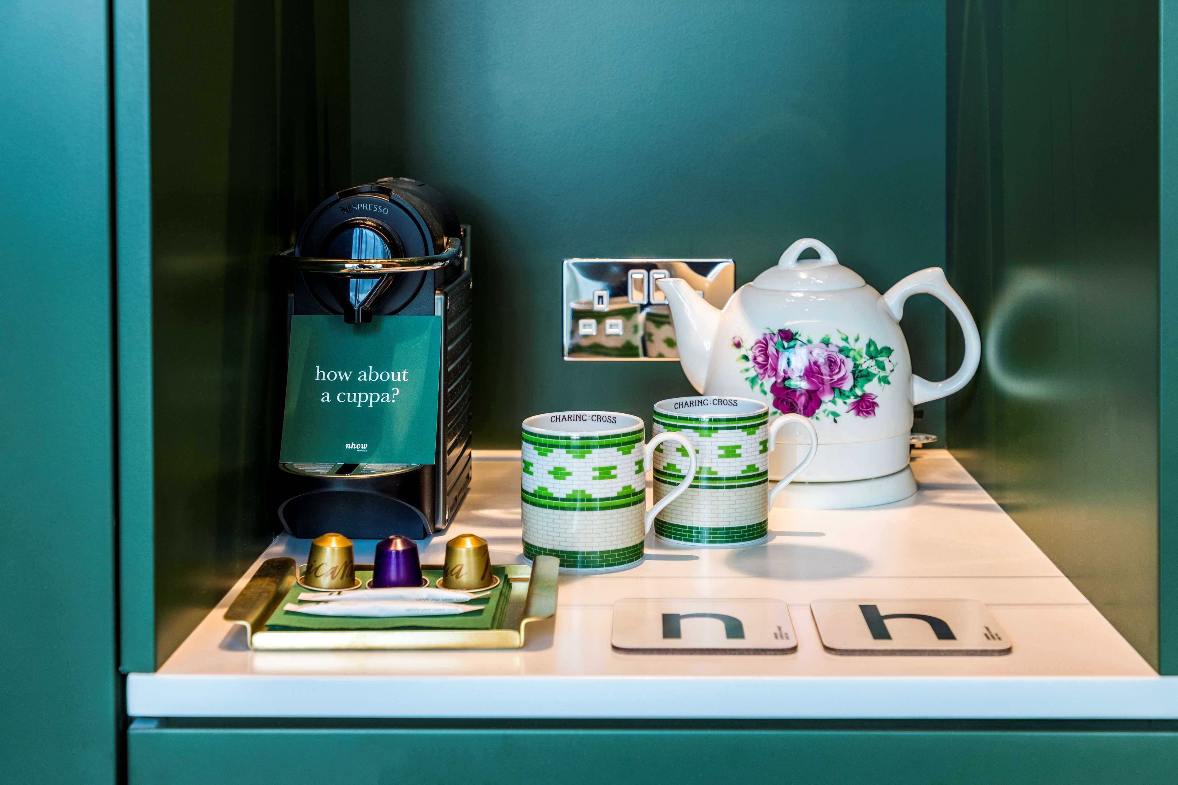 A coffee station with a coffee maker, two mugs, a teapot, and a tray with cups and a spoon.