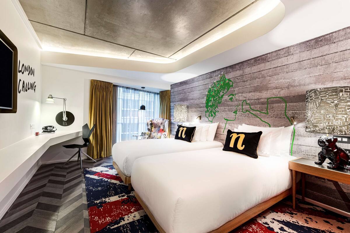 A hotel room with two beds, a desk, a chair, a large window with curtains, and a wall with a green abstract design.