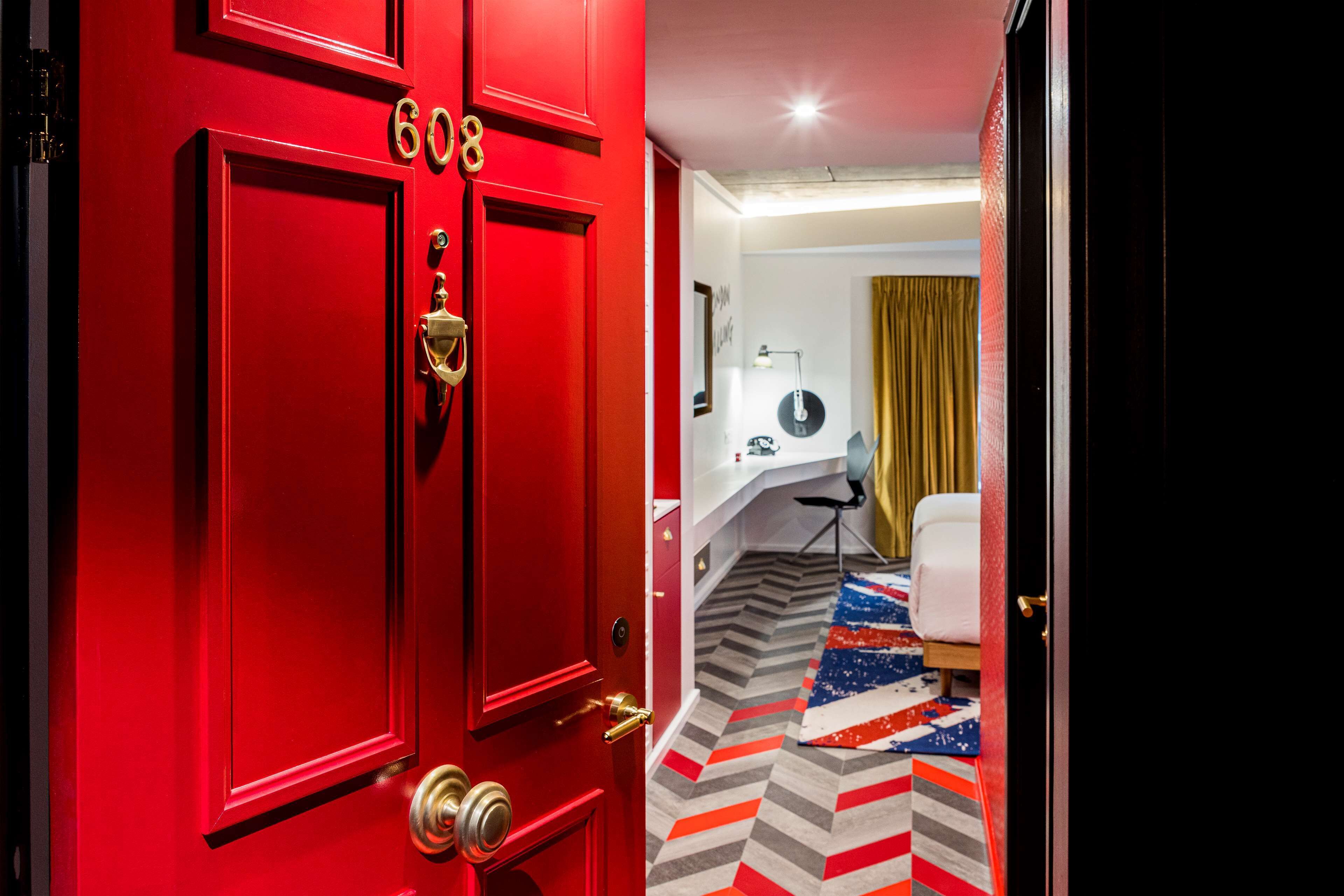 Uma porta vermelha com o número 608, uma secretária branca, uma cadeira, um tapete estampado e uma cortina dourada num quarto.