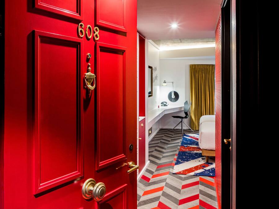 Uma porta vermelha com o número 608, uma secretária branca, uma cadeira, um tapete estampado e uma cortina dourada num quarto.
