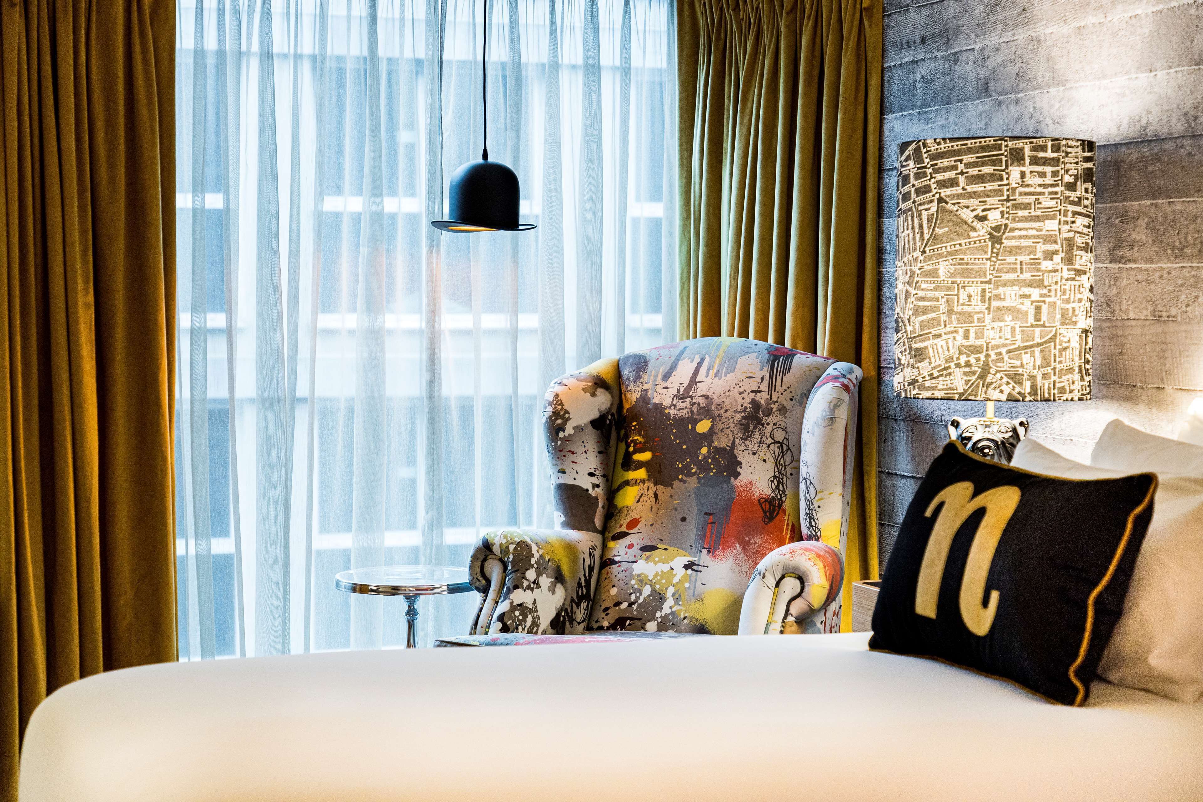 Une chambre d’hôtel avec un lit, un fauteuil coloré, une lampe noire et une fenêtre ornée de rideaux légers.