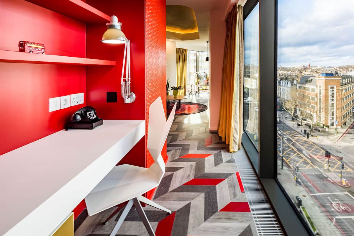A modern hotel room with a red accent wall, a white desk, and a view of the city through large windows.
