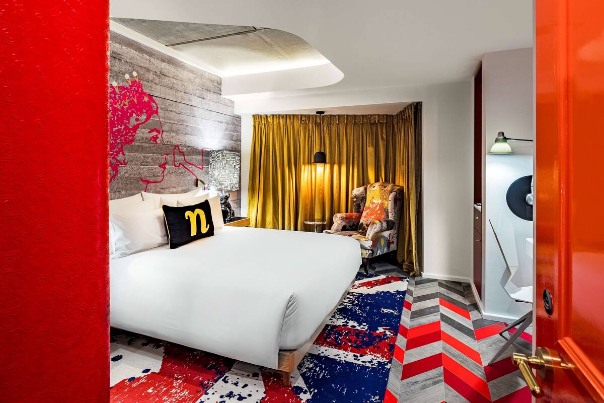 A modern hotel room with a large bed, a red accent wall, a patterned rug, and a golden curtain.