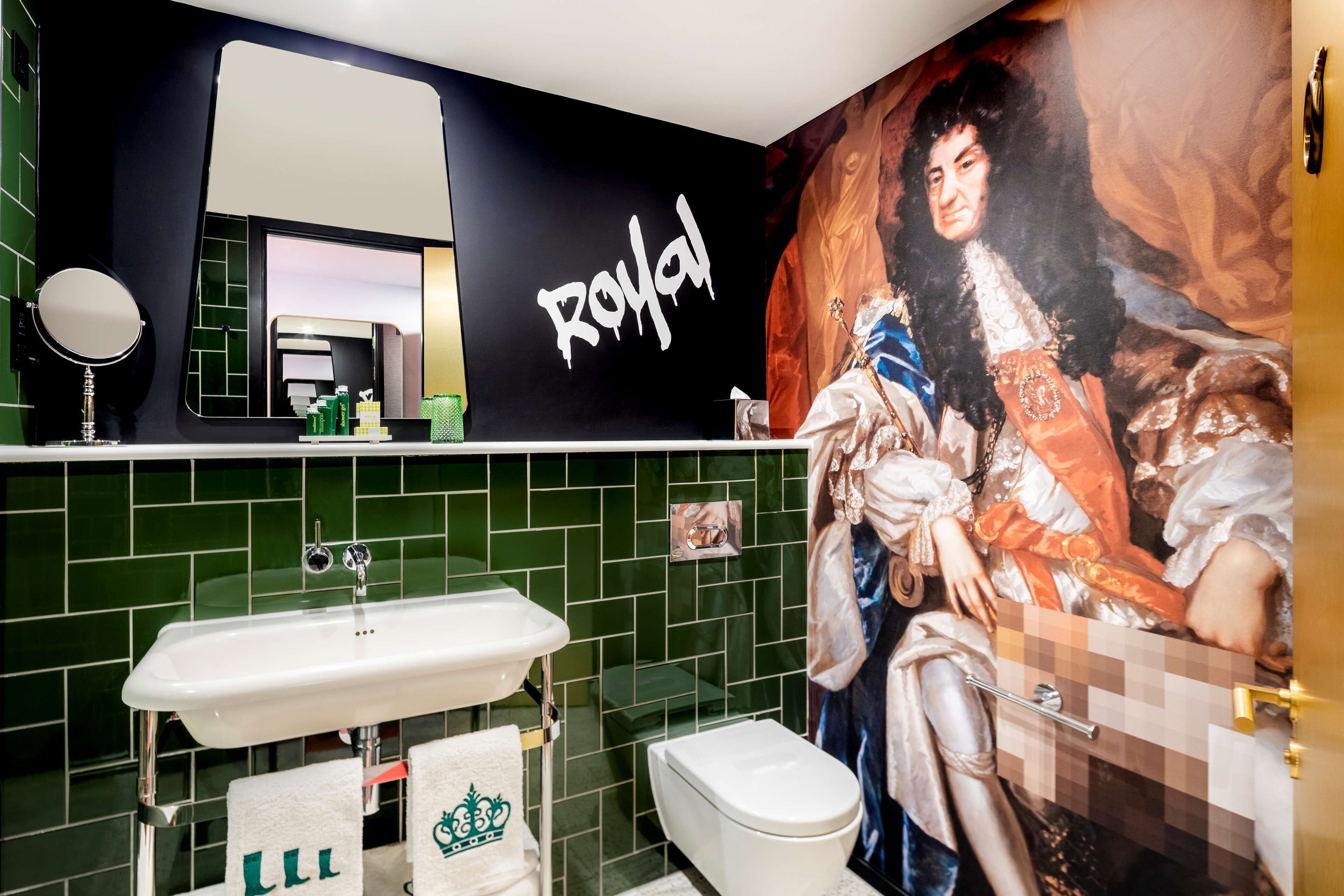 A bathroom with a green tiled wall, a white sink, a mirror, and a toilet. There is a large portrait of a historical figure on the wall.