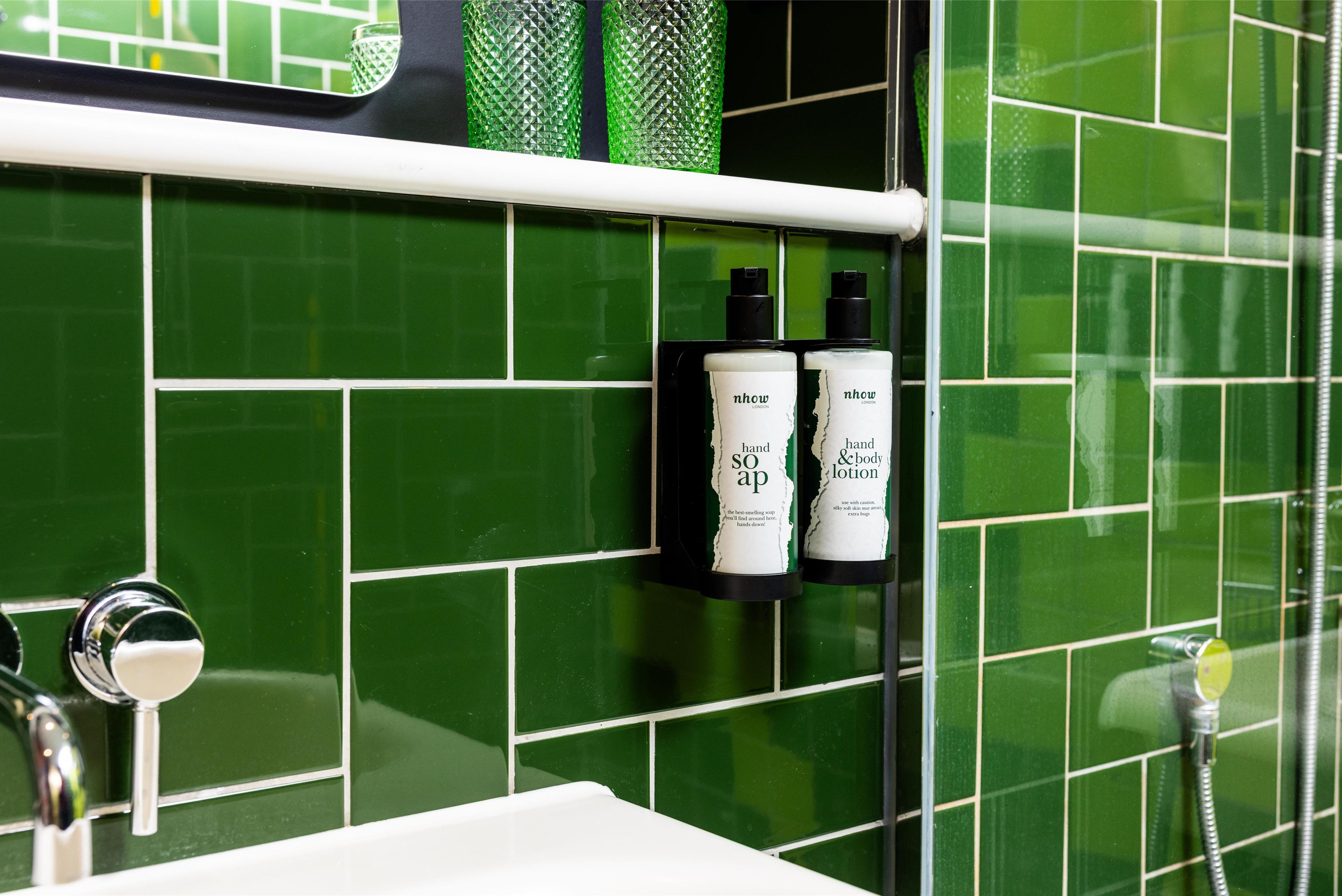 Un bagno con piastrelle verdi, un lavandino bianco e due flaconi di sapone per le mani e crema corpo NHOW su un portaoggetti nero.