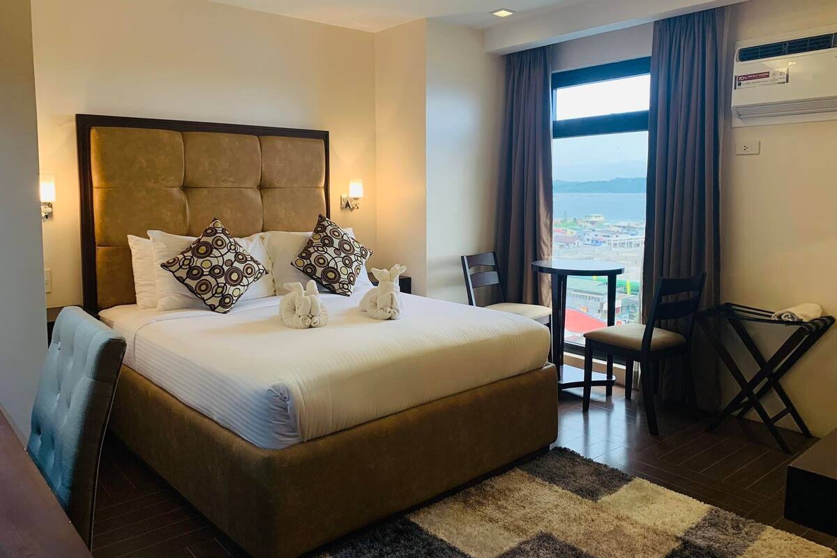 A hotel room with a large bed, two pillows, a small table, a chair, and a view of the city through the window.