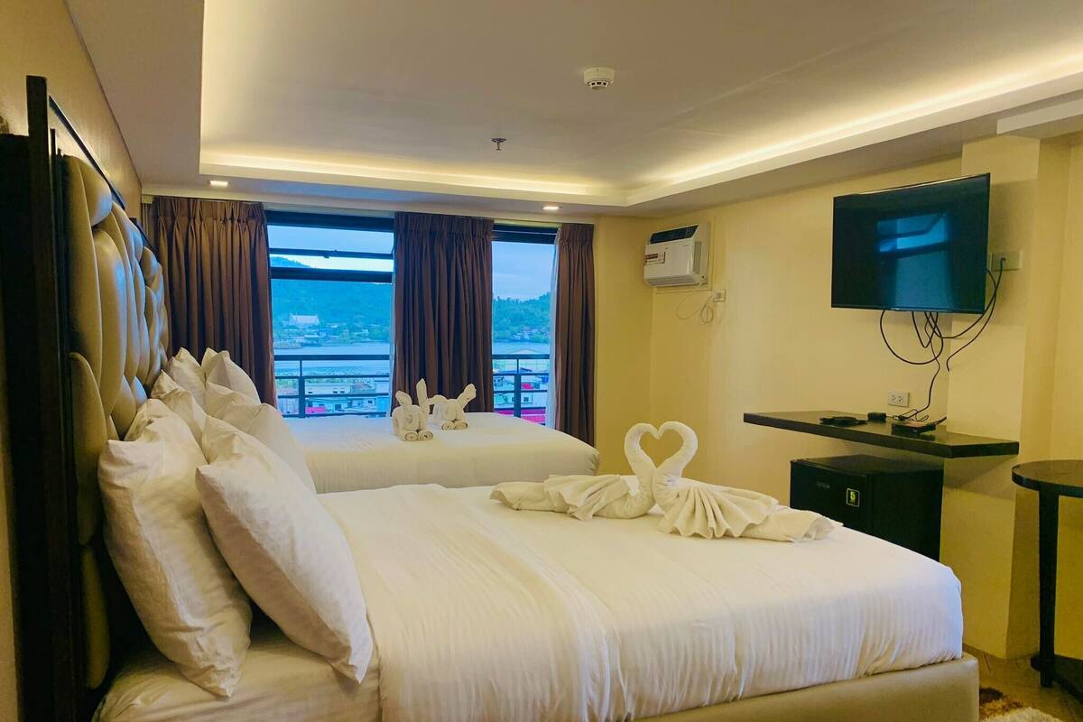 A hotel room with two beds, a TV, a desk, and a view of the sea.