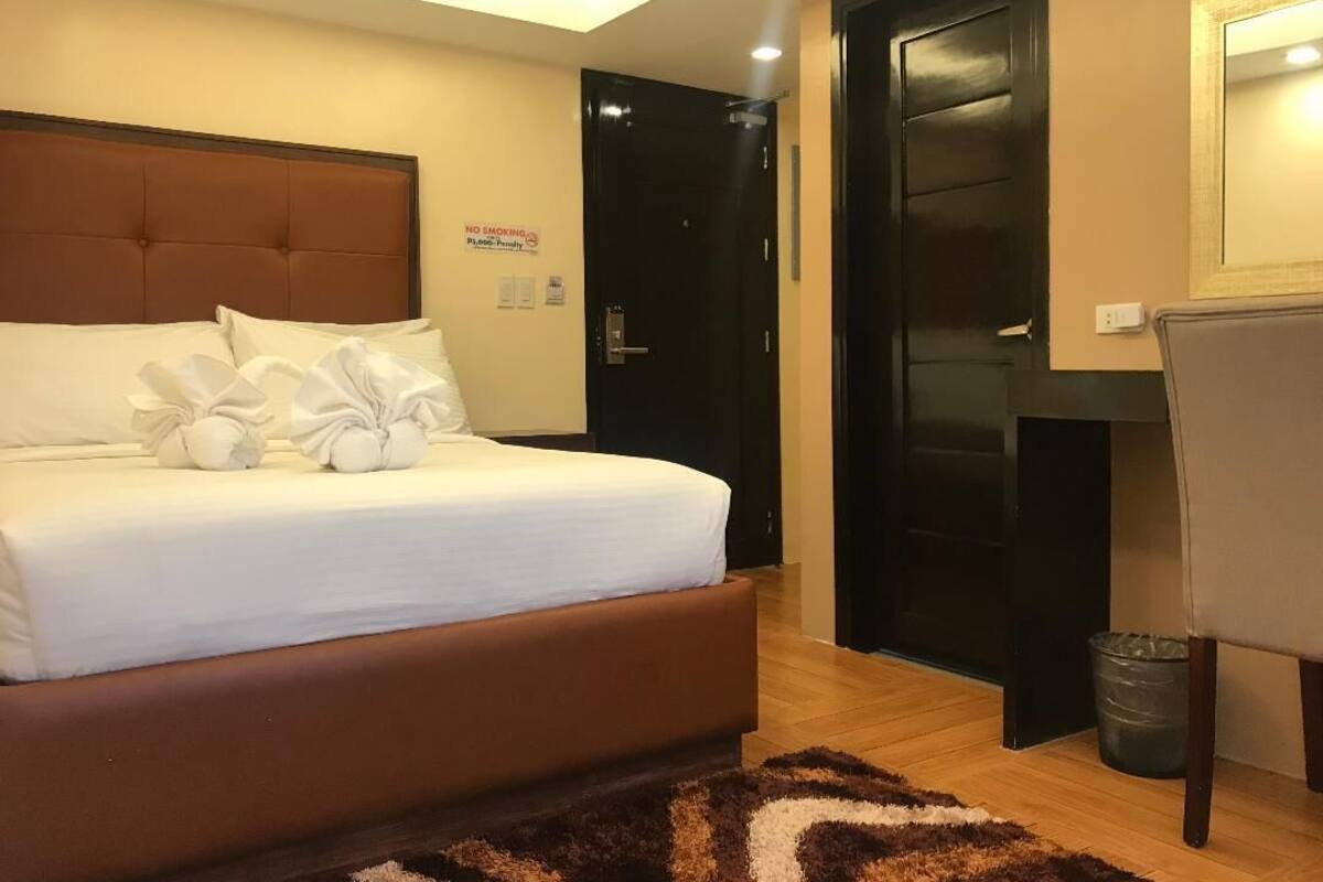 A hotel room with a bed, a door, a chair, and a small trash bin.