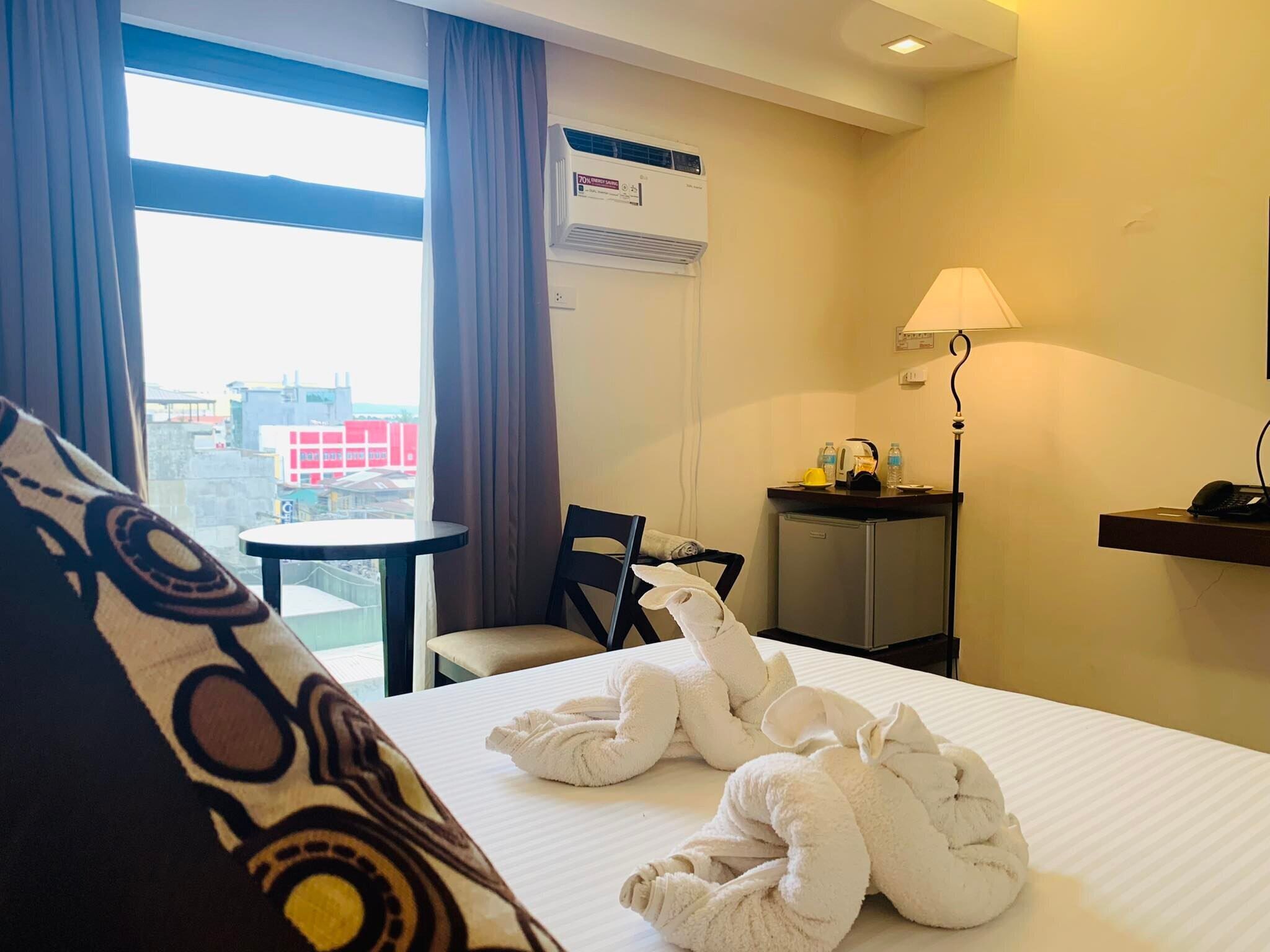 A hotel room with a bed, a desk, a chair, a lamp, and a view of the city through the window.