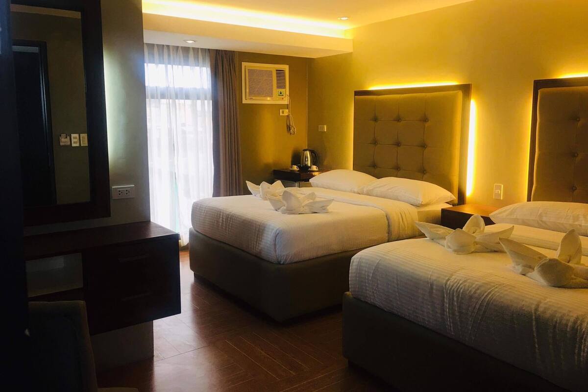 A hotel room with two beds, a large mirror, a bedside table with a lamp, and a window with curtains.