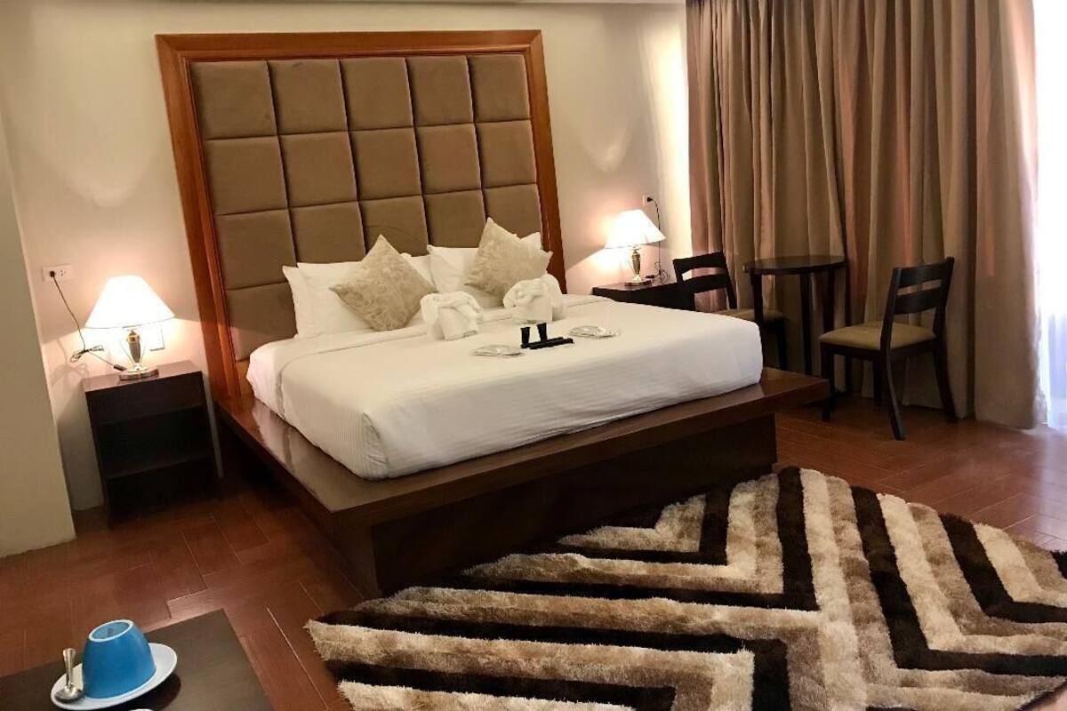 A hotel room with a large bed, two bedside tables with lamps, a chair, a small table, and a patterned rug.