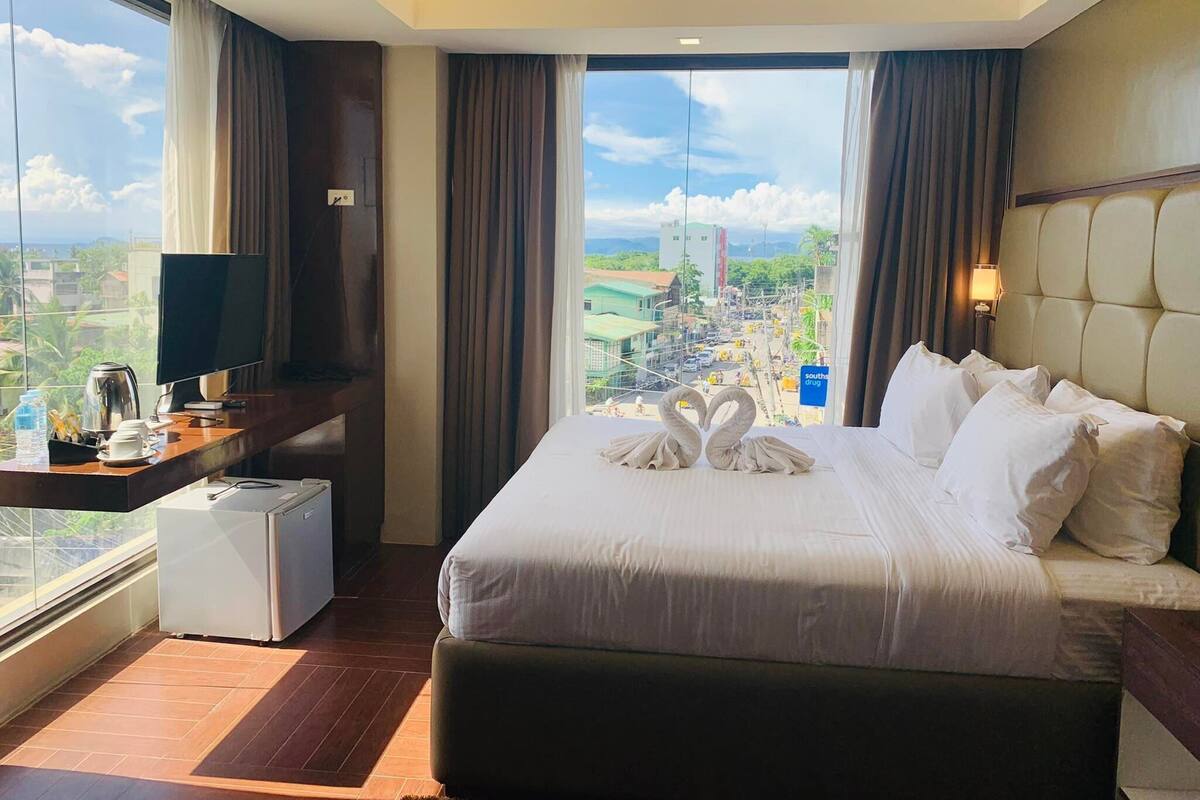 A hotel room with a large bed, a desk with a TV, a mini-fridge, and a view of the city.