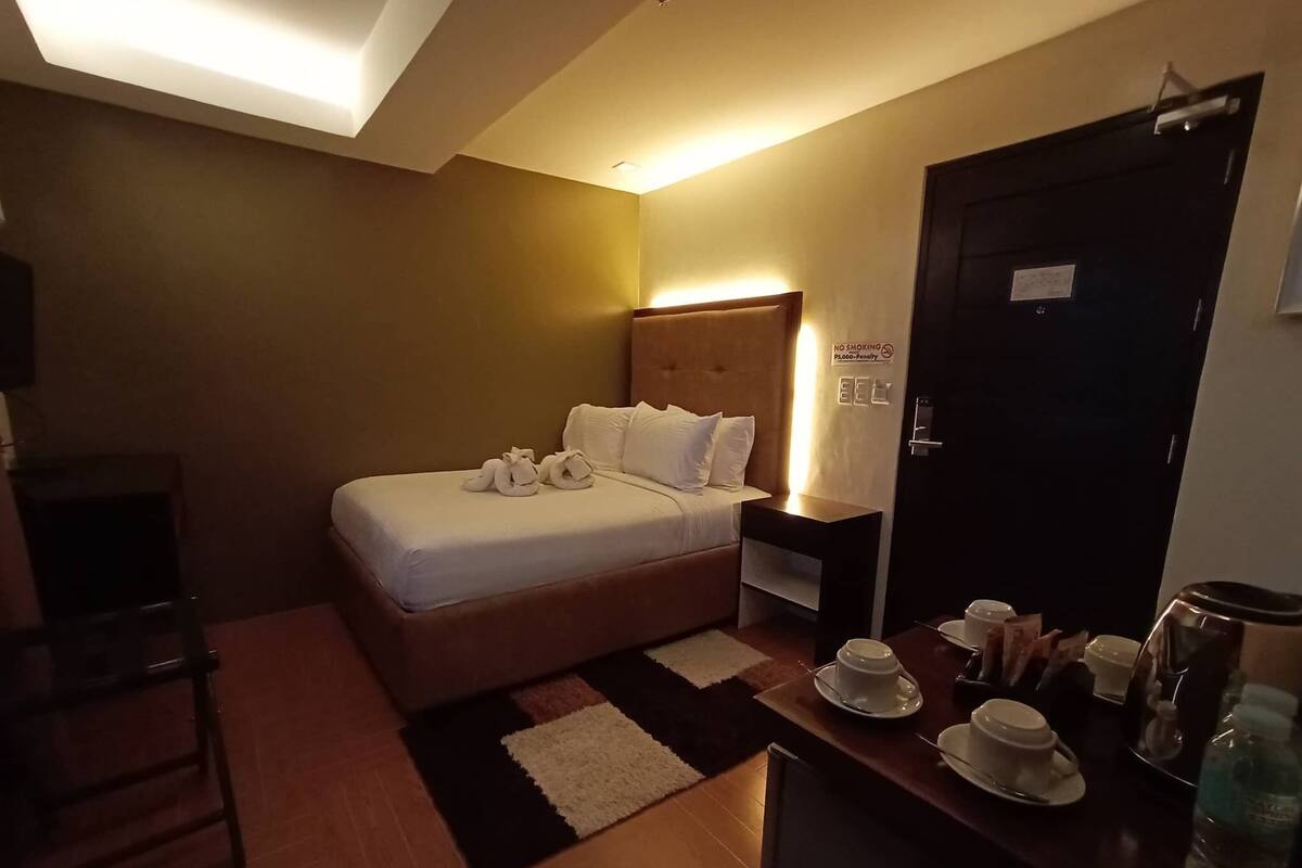 A hotel room with a bed, bedside table with cups and a kettle, a chair, and a door with a no-smoking sign.