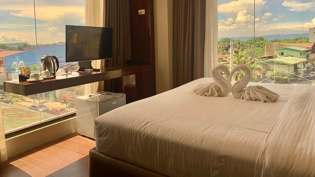 A hotel room with a large bed, a TV on a wooden stand, a view of the cityscape, and a balcony with a table and chairs. - Treasure Island Hotel (Masbate City)