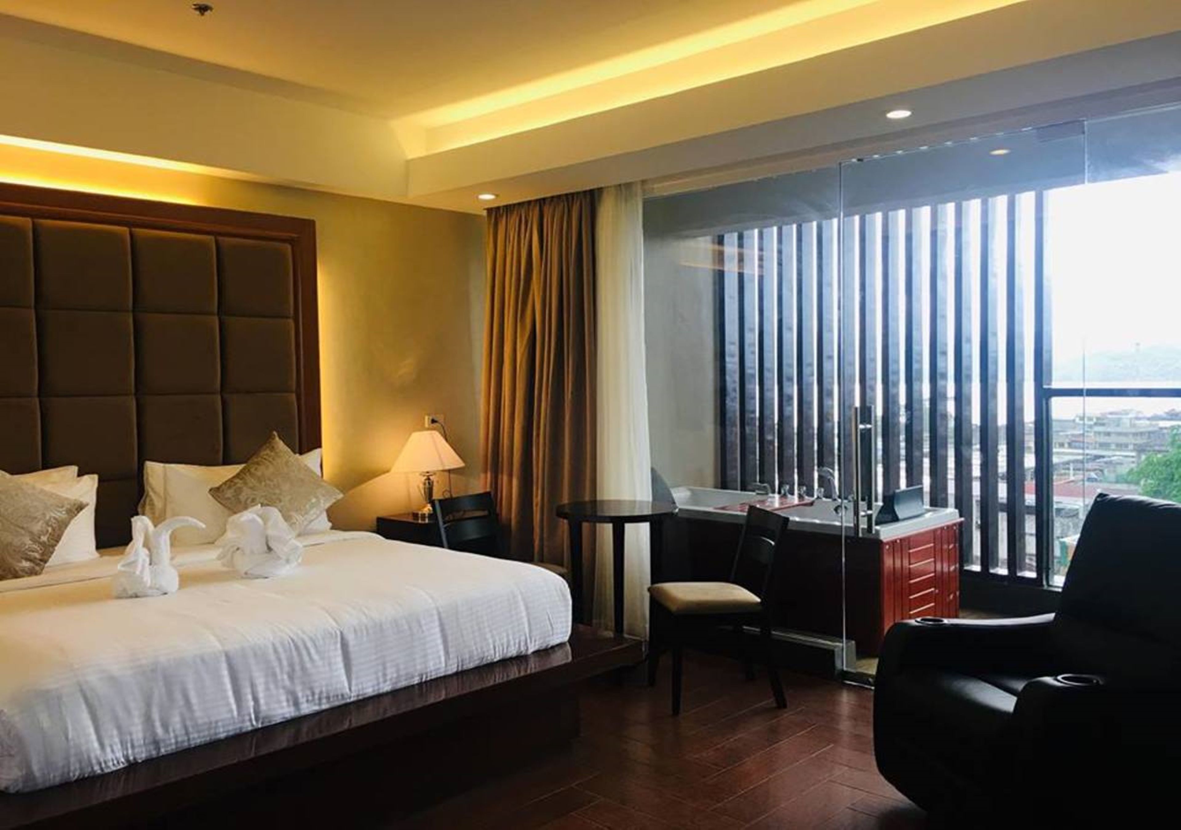 A hotel room with a large bed, a desk, a chair, and a view of the city through large windows.
