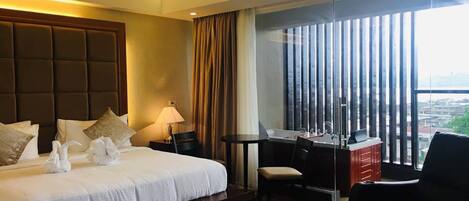 A hotel room with a large bed, a desk, a chair, and a view of the city through large windows.