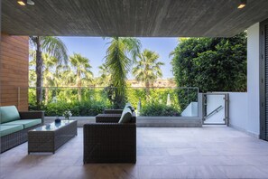 Uma área de estar moderna ao ar livre, com móveis de vime, mesa de centro de vidro e vista para palmeiras e vegetação.