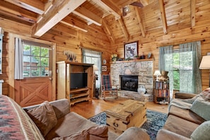 A log cabin interior with a stone fireplace, wooden walls, a television, and a cozy seating area.