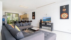 A modern living room with a grey sofa, a coffee table, and a TV on a stand. There is a kitchen area with cabinets and a window. - Villa 3 oceanfront pool villa (Tambon Patong)