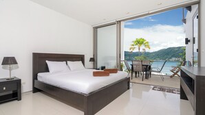 A modern bedroom with a large bed, a nightstand, a desk with a chair, and a balcony with a view of the sea and mountains. - Villa 3 oceanfront pool villa (Tambon Patong)