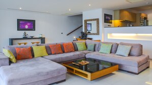 A modern living room with a sectional sofa, a glass coffee table, and a kitchen area in the background. - Villa 0. oceanfront pool villa (Patong)