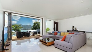 A modern living room with a large window, a grey sofa, a coffee table, and a dining area with chairs. - Villa 0. oceanfront pool villa (Patong)