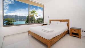 A bedroom with a large window offering a scenic view of a coastal landscape, a wooden bed, a bedside table, and a nightstand. - Villa 0. oceanfront pool villa (Patong)