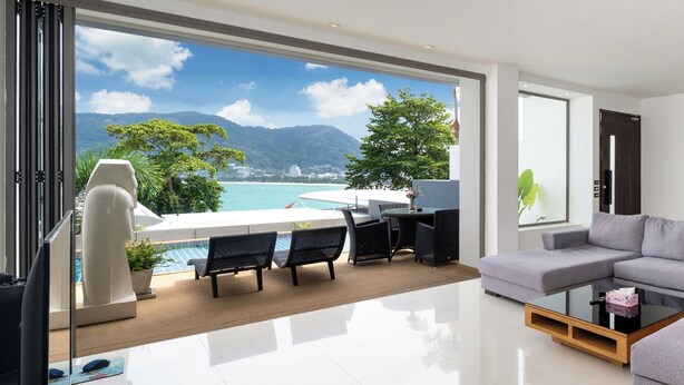 A modern living room with a large window overlooking a pool and mountains. There is a sofa, a coffee table, and a desk with a chair. - Villa 0. oceanfront pool villa (Patong)