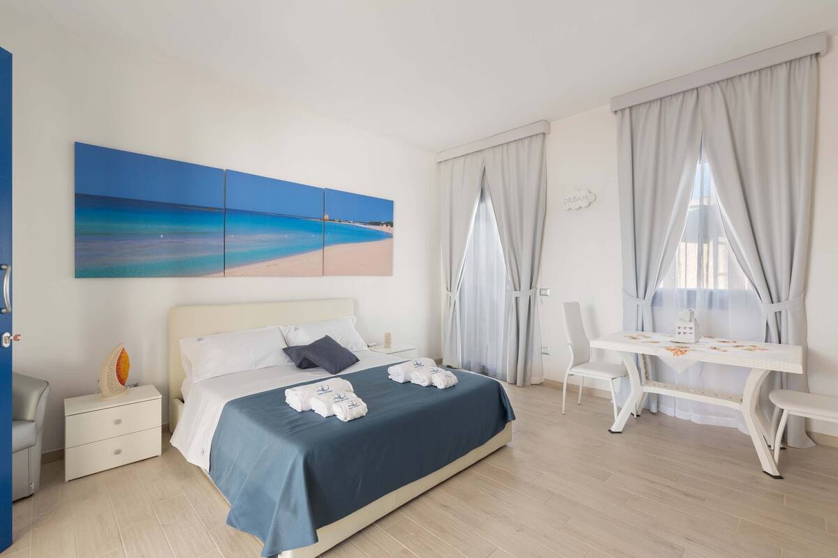 Ein Hotelzimmer mit einem Bett, einem Schreibtisch und einem Sessel. An der Wand hängt ein Gemälde und ein Fenster mit Vorhängen.