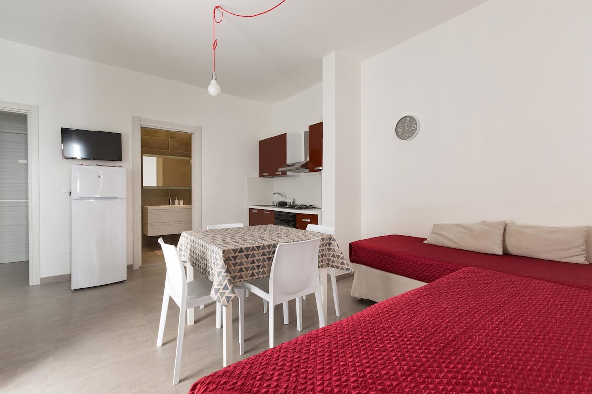 A compact room with a bed, a dining table with chairs, a kitchenette, a TV, and a clock on the wall.