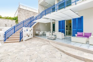 A modern outdoor living area with a stone floor, white furniture, blue railings, and a covered seating area. - Villa Syrmata (Porto Cesareo)