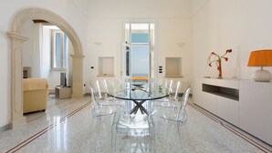 A modern dining area with a glass table and chairs, a beige sofa, and a view of the sea through an open doorway. - Villa Dorella (Castrignano del Capo)