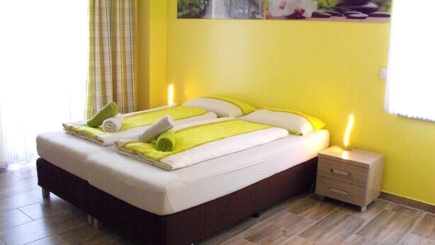 Ein ordentlich bezogenes Bett mit gelb-weißer Bettwäsche, ein Nachttisch mit Lampe und ein Fenster mit Vorhängen.