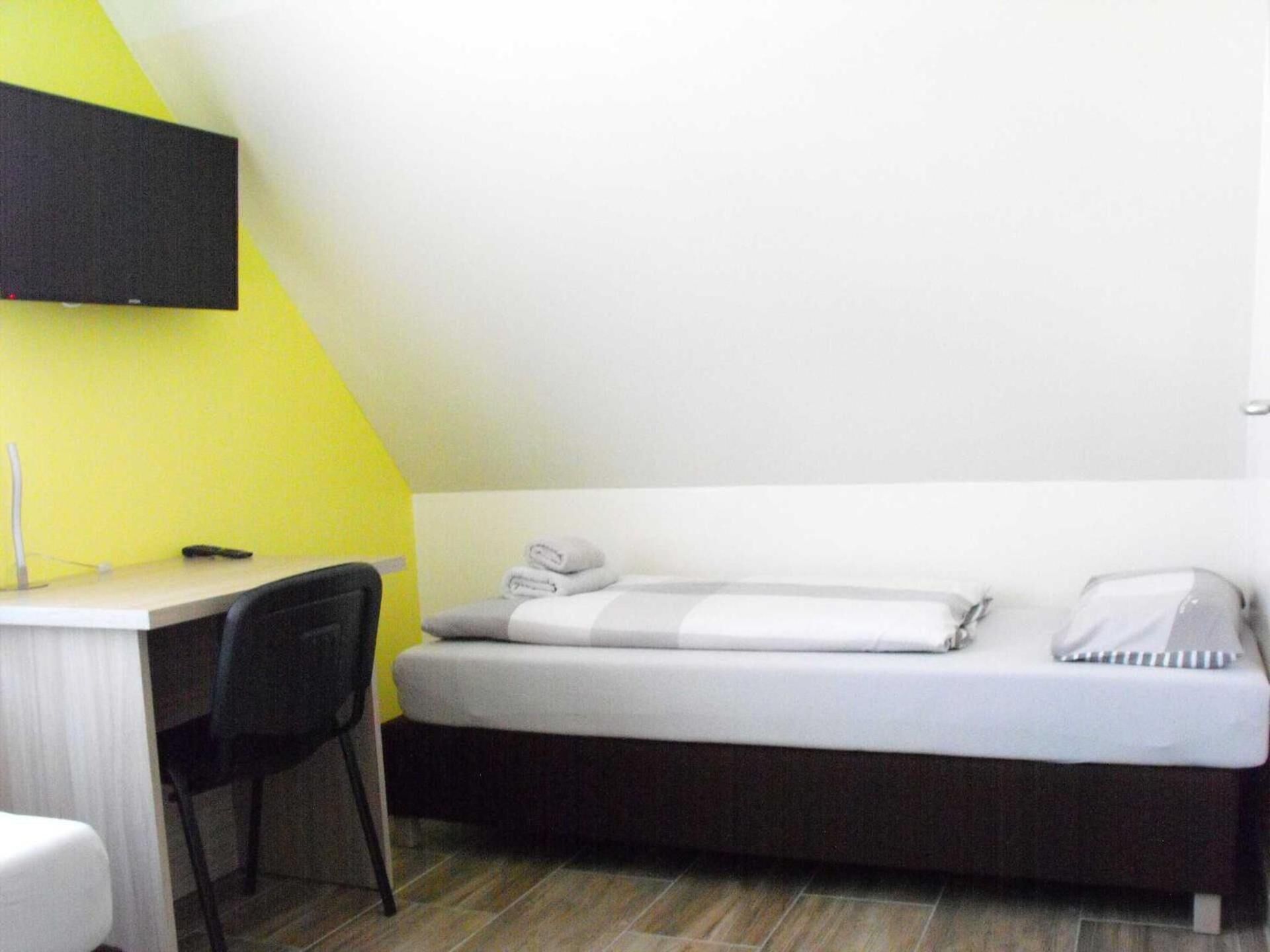 A small, single-bed room with a desk and chair, a flat-screen TV mounted on the wall, and a yellow accent wall.