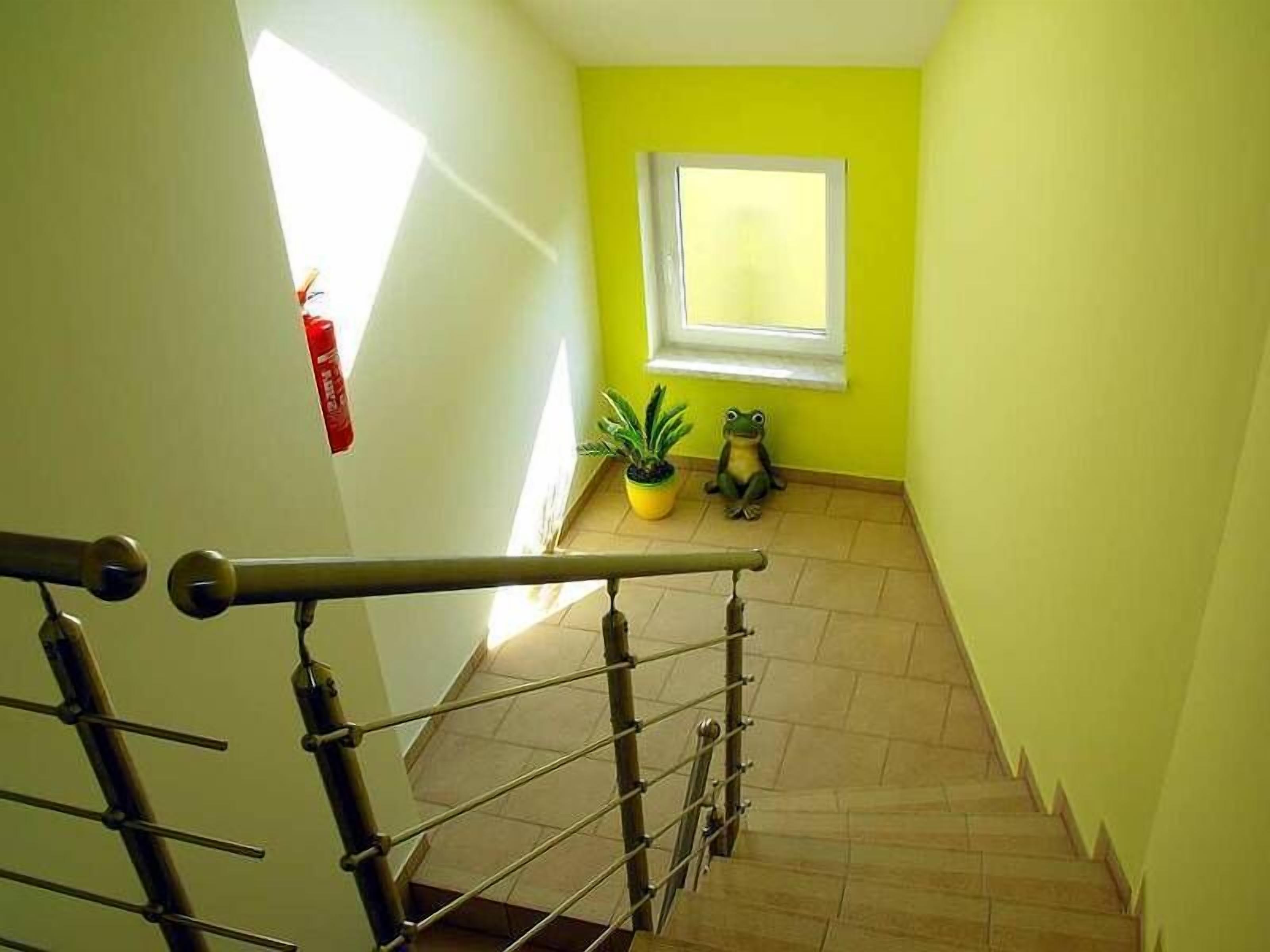 Una escalera con barandilla de metal, un extintor en la pared, una ventana con vistas y una planta en maceta en el suelo.