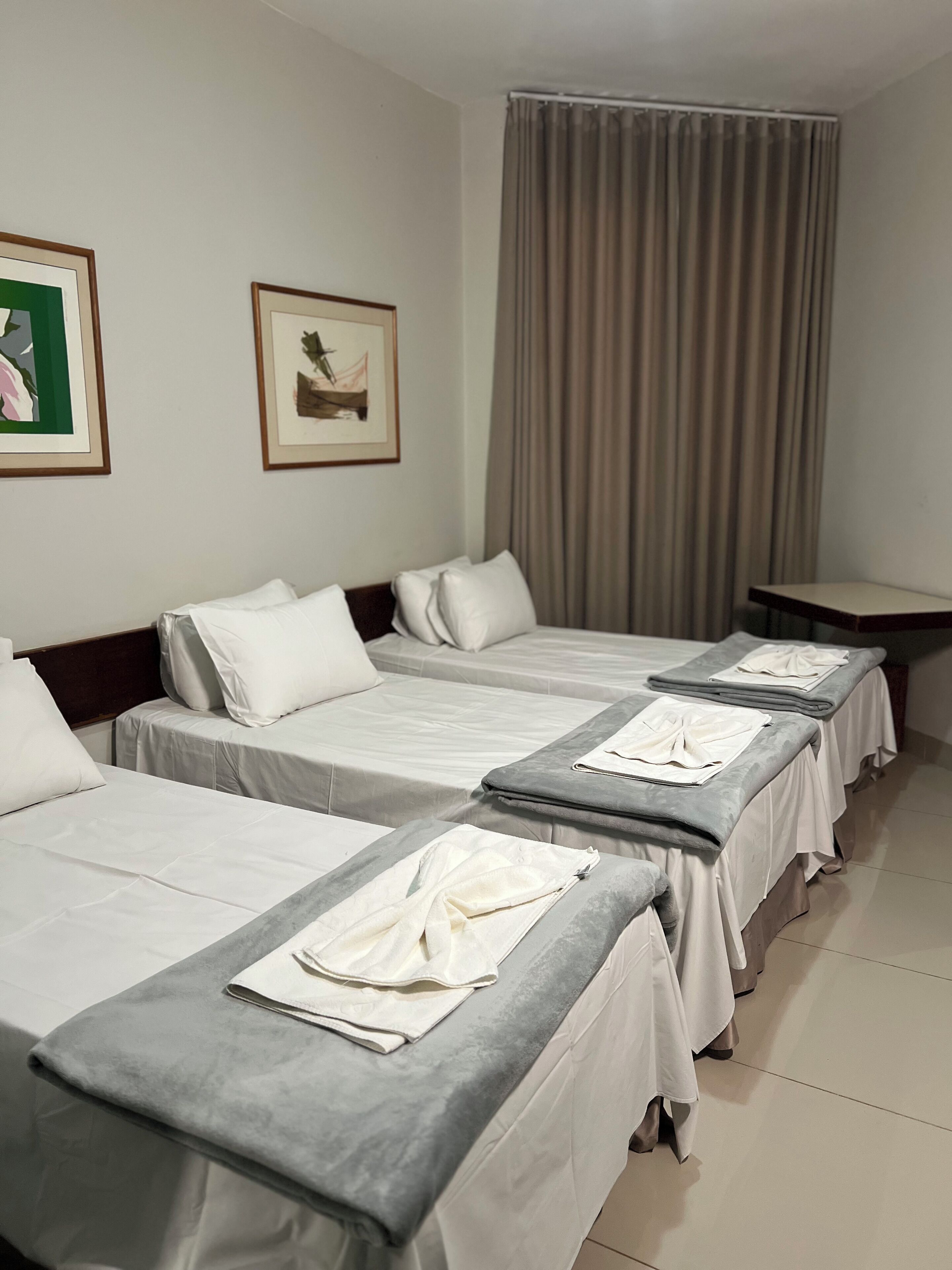 A hotel room with four beds arranged in a row, each with white bedding and grey headboards. There are two framed pictures on the wall, a window with curtains, and a small table in the corner.