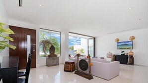 A modern living room with a white sofa, a dining table with chairs, a wooden statue, a decorative wheel, and a TV mounted on the wall. - Atika Villa 1 oceanfront pool villa (Patong)