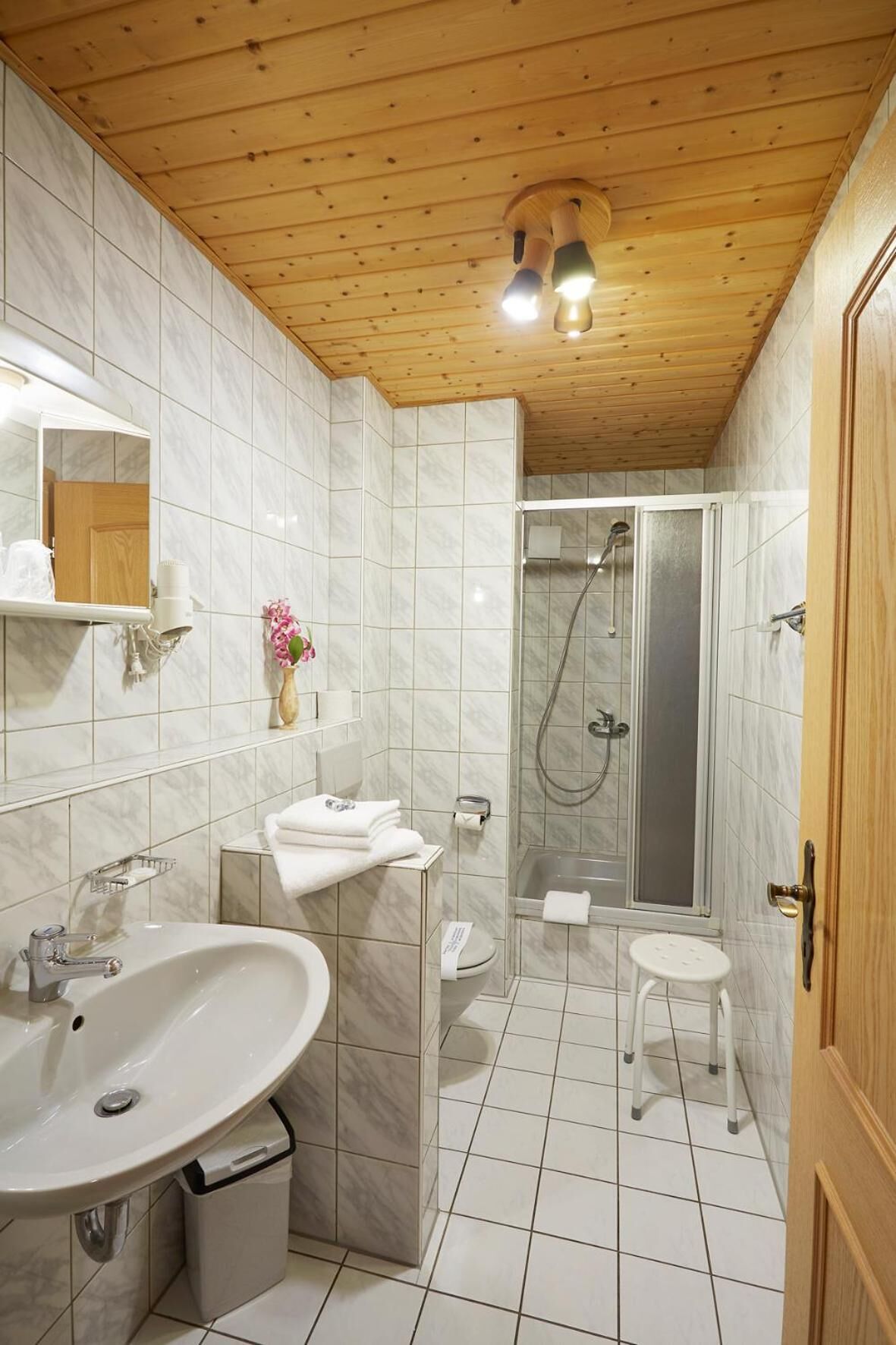 Ein Badezimmer mit Holzboden, einem Waschbecken, einer Toilette, einer Dusche und einem kleinen Hocker.