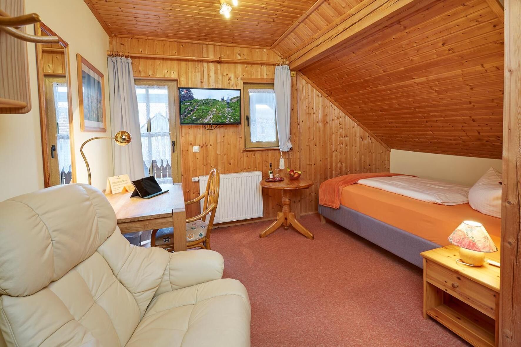 A cozy wooden room with a bed, a desk with a laptop, a chair, and a TV mounted on the wall.
