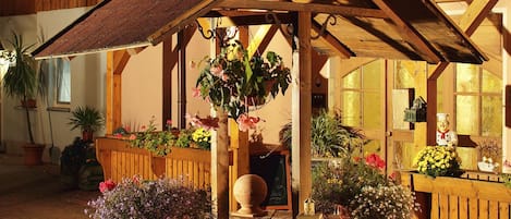 A rustic wooden building with a stone fountain, potted plants, and string lights.