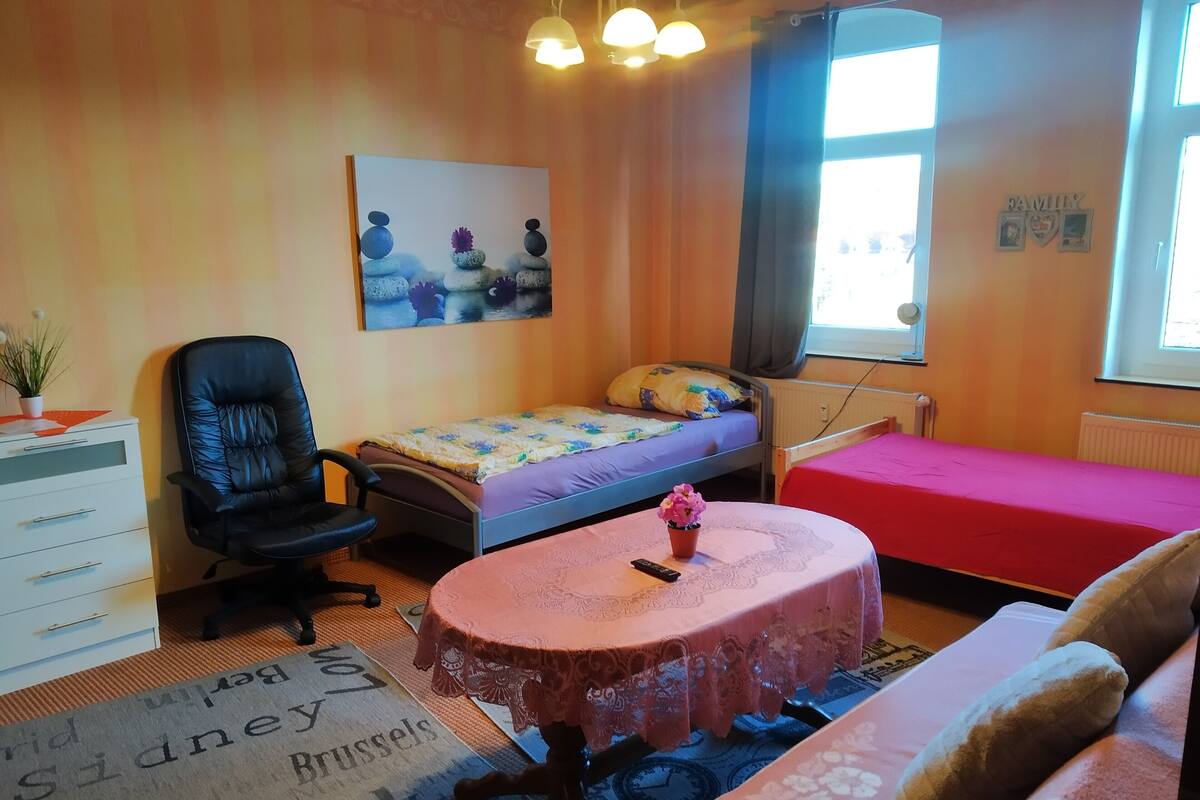 A bedroom with a bed, a chair, a small table with a tablecloth, a painting on the wall, and a window.